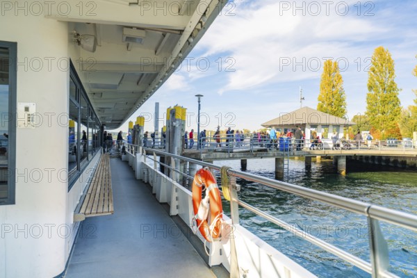 Harbour with people on a sunny day, boats in water, trees in shades of yellow and orange, Überlingen, Lake Constance, Germany