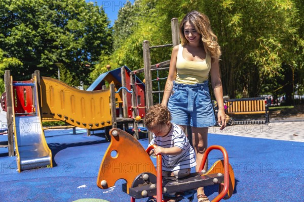 Young mother assisting her son while playing on a rocking horse in a colorful playground on a sunny summer day