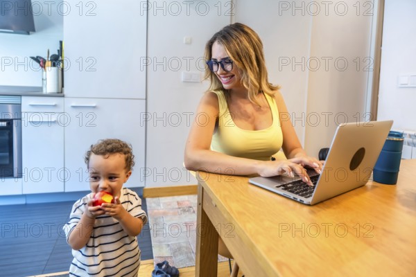 Woman working remotely on a laptop in her kitchen while taking care of her baby son eating an apple