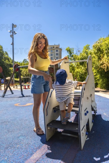 Happy mother assisting her toddler son while he's climbing stairs on a playground structure, enjoying a sunny day at the park