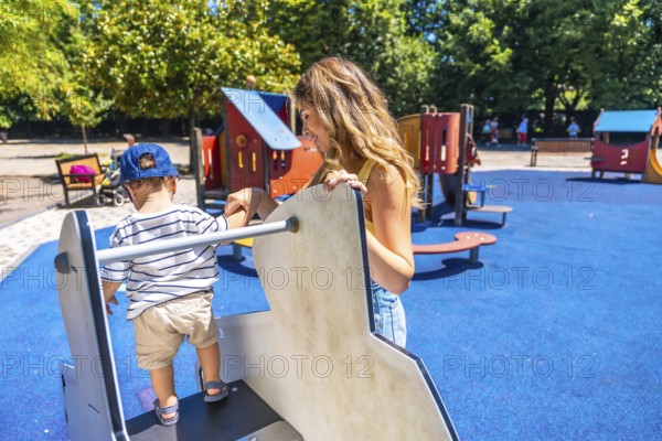Young mother assisting her little son while he is carefully climbing on a playground structure, enjoying a sunny day at the park