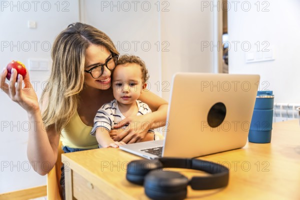 Mother holding an apple while hugging her son, both enjoying a moment together, watching something entertaining on the laptop at home