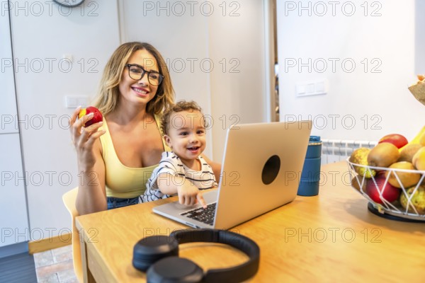 Mother holding an apple while son focused on laptop in kitchen, surrounded by a fruit basket, enjoying a healthy and happy moment together