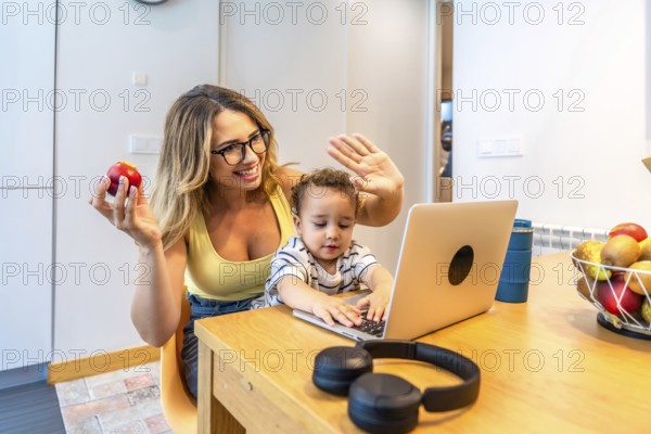 Mother and son waving joyfully at the computer during a video call while enjoying breakfast together in the cozy kitchen