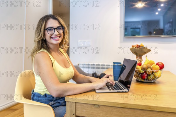 Smiling woman using laptop at kitchen table, working remotely with fruit basket and headphones nearby