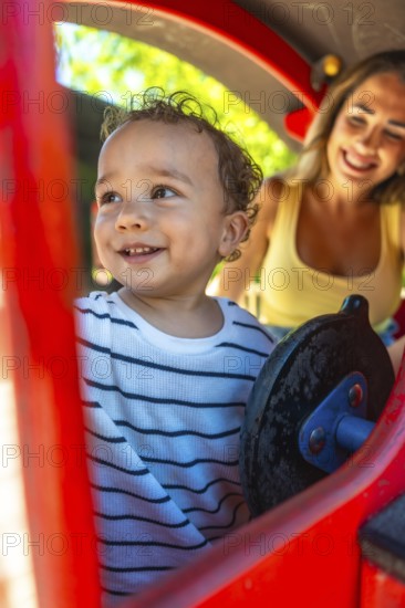 Smiling toddler playing joyfully on a playground slide while mother watches nearby, creating moments of fun and togetherness in the sun
