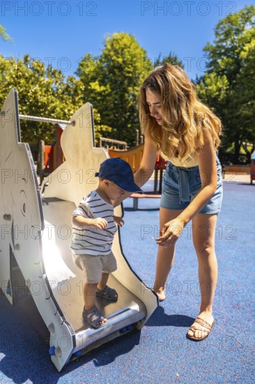 Mom carefully assisting her little boy while he's learning to use the slide at a children's playground on a sunny summer day