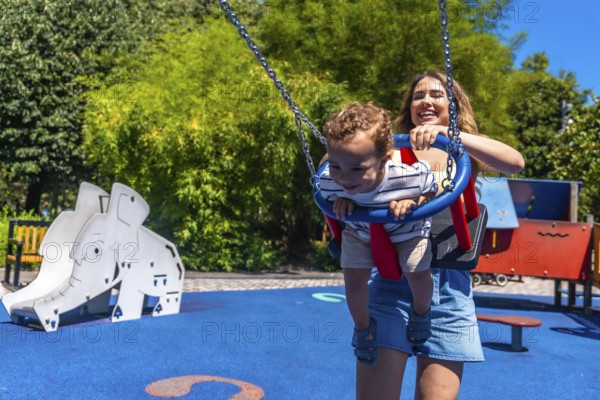 Young mother having fun with her baby son, pushing him on a swing in a colorful playground during a sunny summer day