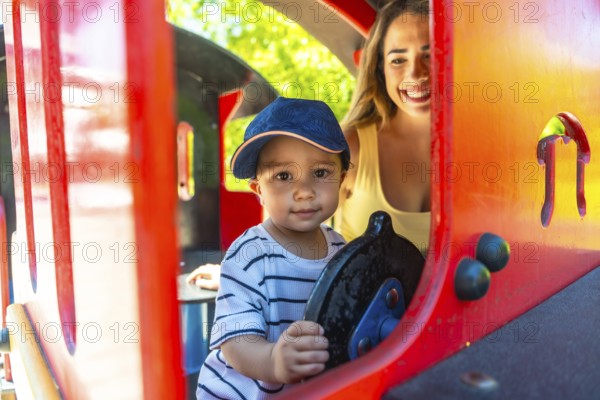 Happy toddler having fun driving a toy train at the playground while his mother watches him with a smile