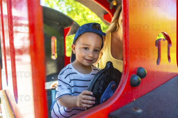 Little boy having fun driving a toy train at the playground, with his mother supervising in the background, enjoying a sunny day outdoors