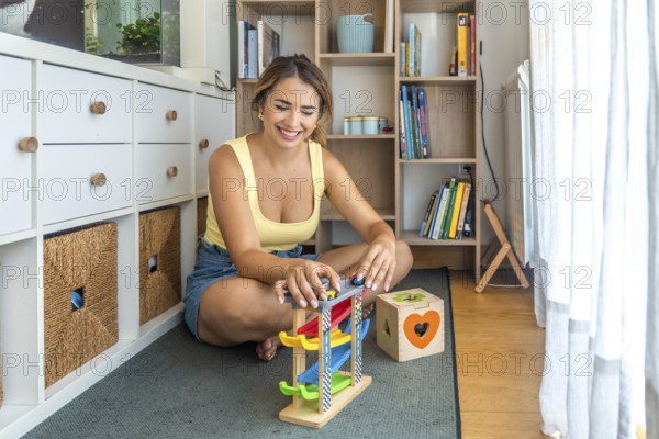 Young woman playing with a toy car ramp and smiling while sitting on a play mat in a child's playroom