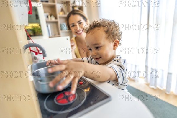 Happy toddler boy having fun playing with a toy kitchen while his mother watches over him in a bright and airy home