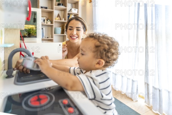 Smiling mother looking at her little son having fun playing with a toy kitchen in a modern, bright living room