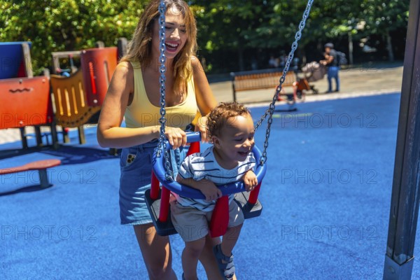 Happy mother enjoying quality time with her toddler son, playfully pushing him on a swing at the vibrant playground on a sunny day