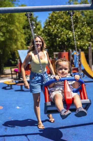 Young child enjoying a swing at the playground with their mother looking on, having fun on a summer day