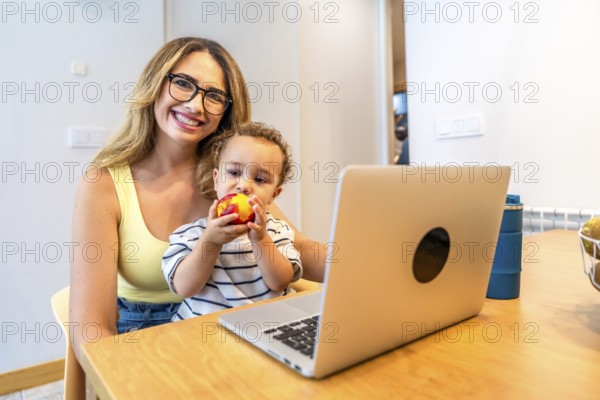 Mother holding her son while enjoying a nectarine and working on a laptop in the bright kitchen, fostering connection and togetherness