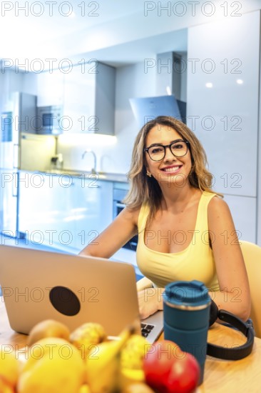 Young woman working on laptop in modern kitchen with fresh fruits, promoting healthy work life balance and remote work opportunities
