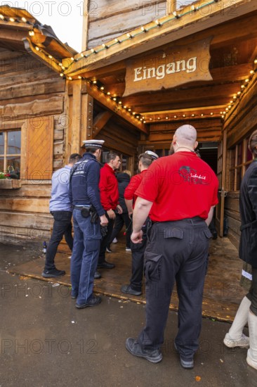 People stand in front of a wooden entrance, some wearing red shirts. A security-conscious atmosphere in a traditional setting, Cannstatter Wasen folk festival, Sonja Merz marquee, Stuttgart, Germany