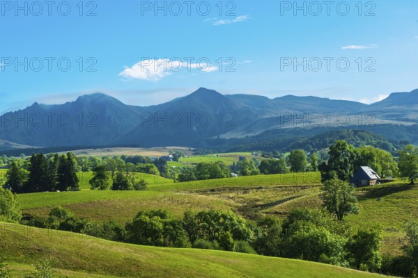 Lush green fields with a solitary house, set against majestic mountains and a bright sky, Massif of Sancy in Auvergne Volcanoes Regional Park. Puy de Dome. Auvergne Rhone Alpes. France