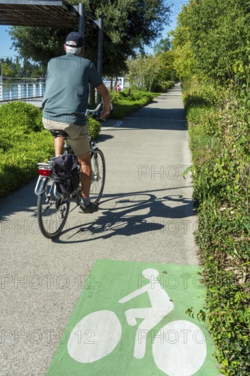 A person rides a bicycle on a smooth pathway lined with trees and shrubs. The sun shines bright, creating a pleasant atmosphere for outdoor activities. Vichy. Allier. Auvergne Rhone Alpes. France