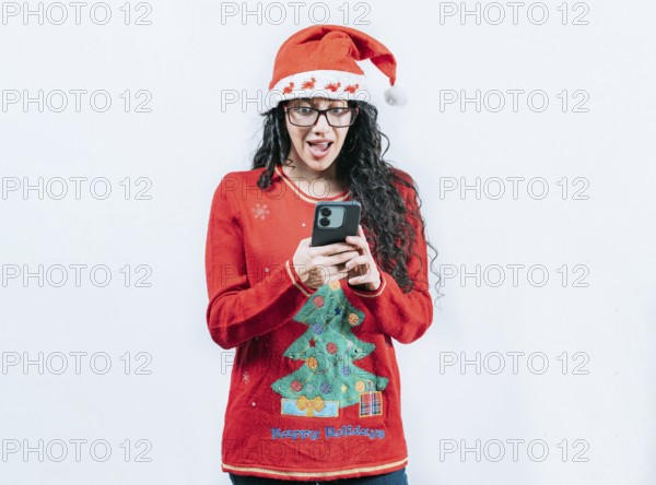Amazed girl in Christmas hat looking at phone isolated. Surprised girl in Christmas sweater looking at cell phone screen, isolated