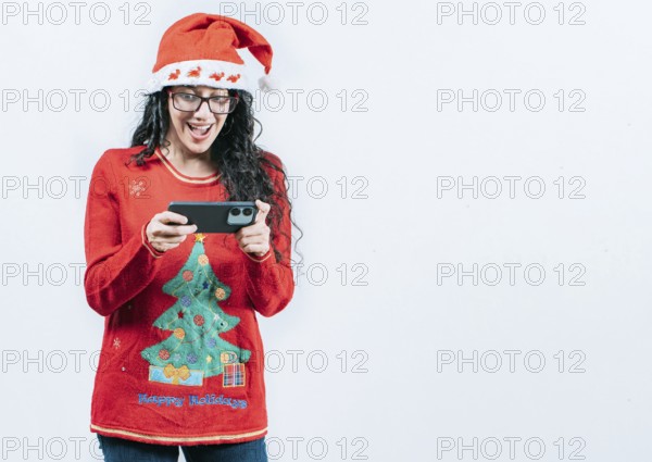 Surprised girl in Christmas sweater looking at cell phone screen, isolated. Surprised girl in Christmas hat looking at phone isolated