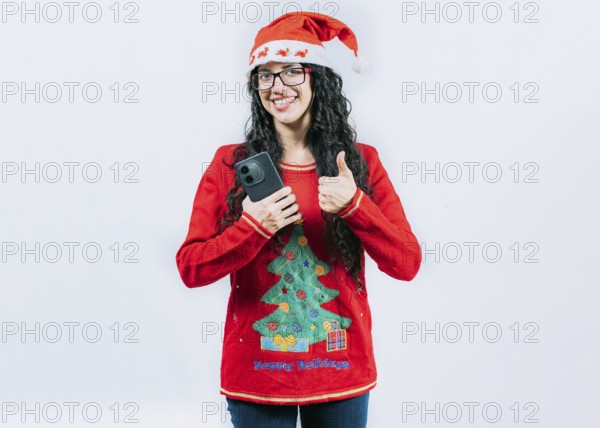 Smiling girl in Christmas hat holding phone with thumb up isolated. Positive young woman in Christmas hat holding smartphone gesturing ok