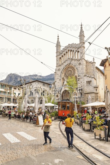 Waiters walking on a square with outdoor restaurants and an old vintage tram driving across the square with lot of tourists, Soller, Mallorca, Spain