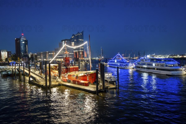 View of illuminated ships on the Elbe promenade with restaurant Das Feuerschiff LV 13, City Sporthafen, behind Speicherstadt with Elbphilharmonie and Columbus Haus, blue hour, Hamburg, Germany