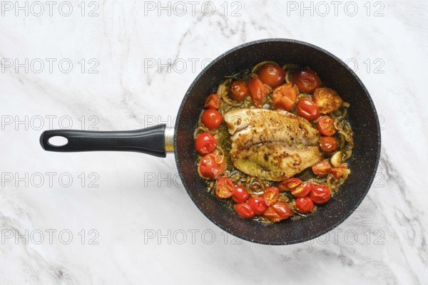 A frying pan holds a seasoned fish fillet surrounded by bright cherry tomatoes. The scene captures a fresh, healthy meal preparation on a modern kitchen counter with marble design