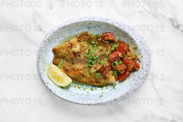 A perfectly cooked white fish fillet is topped with sautéed tomatoes and fresh herbs. A lemon wedge adds a touch of freshness to this colorful dish, presented on a stylish plate