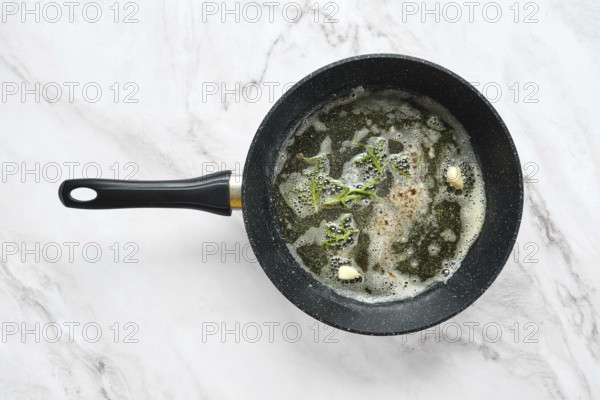 A frying pan is on a marble surface, filled with bubbling butter, fresh garlic and frosemary. Infusing the oil with aroma