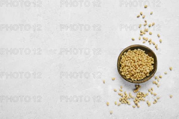 A wooden bowl filled with golden pine nuts on a light gray textured surface. Some nuts are scattered around the bowl, creating a natural and organic feel to the scene