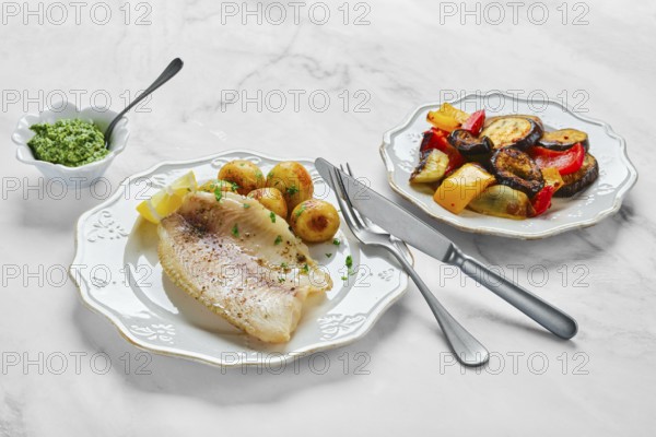 A beautifully arranged meal features tender fish fillet alongside roasted colorful vegetables and baby potatoes. Garnished with herbs, the dishes are set on white marble