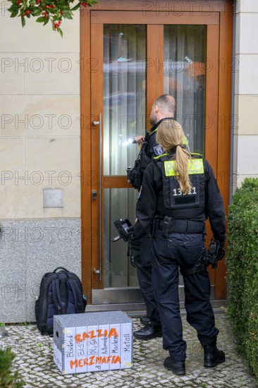 The New Generation symbolically organises a house search at the Family Business and Politics Foundation. Two police officers stand in front of an entrance covered with posters and boxes labelled Razzia Merzmafia: Blackrock, BASF, Öl, Gas, Automobil, Masken, Metall lie in front of the building, police officers enter. Berlin, 10.10.2025