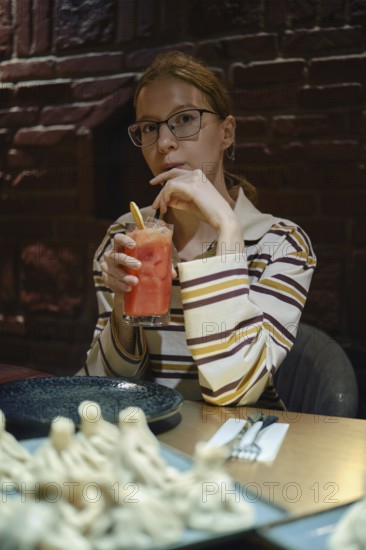 A young woman sits at a wooden table in a warm, dimly lit restaurant. She holds a colorful drink with a straw, gazing thoughtfully