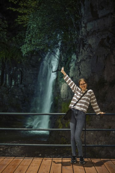 A cheerful young woman stands on a wooden walkway behind a railing, posing joyfully with one arm raised