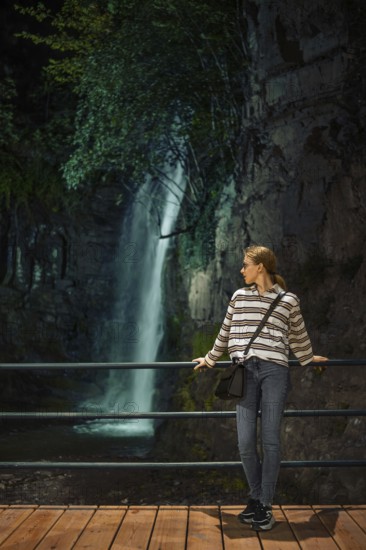 A young woman enjoys a quiet moment by a waterfall in a lush, natural setting during the evening hours. The scene is illuminated by soft evening light, enhancing the tranquil atmosphere