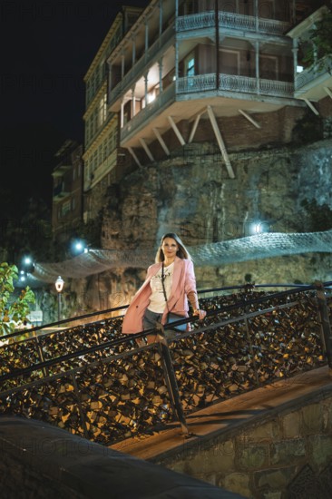 A woman stands on a decorative bridge at night. The backdrop showcases illuminated historic buildings and a rocky cliff, creating a vibrant cityscape atmosphere