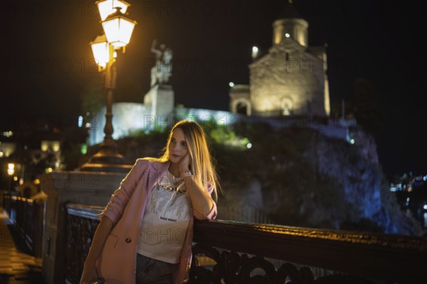 A woman leans against a railing at night, dressed casually with a light jacket. Behind her, a historic castle is illuminated by soft lights, adding charm to the evening atmosphere