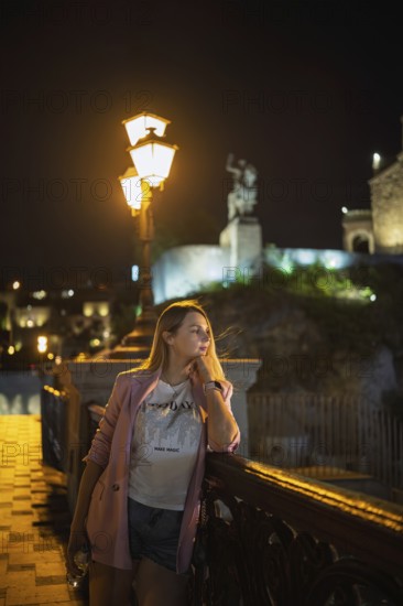A woman stands by a decorative railing of the bridge, admiring the night scenery. Dim lights illuminate the surrounding historical buildings in the city center