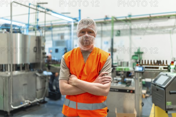 Worker standing proudly with crossed arms, wearing a safety vest and hairnet in a clean, automated beer bottling plant, overseeing the manufacturing process