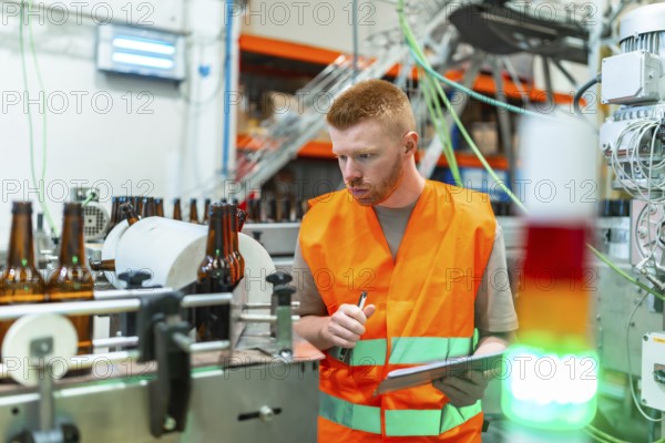 Young man in safety vest checking beer bottles on a factory conveyor belt, performing quality control during the industrial bottling and packaging process