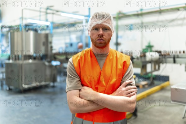 Male worker standing with crossed arms, wearing a hair net and a safety vest, overseeing operations in a food and beverage production facility with machinery in the background