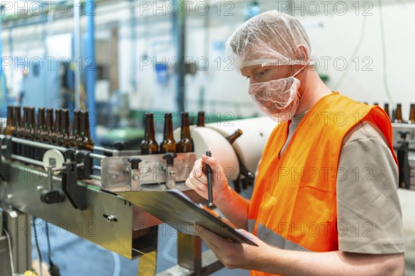 Worker in a hairnet and face mask checking clipboard records while brown beer bottles move along a production line conveyor belt in a beverage factory