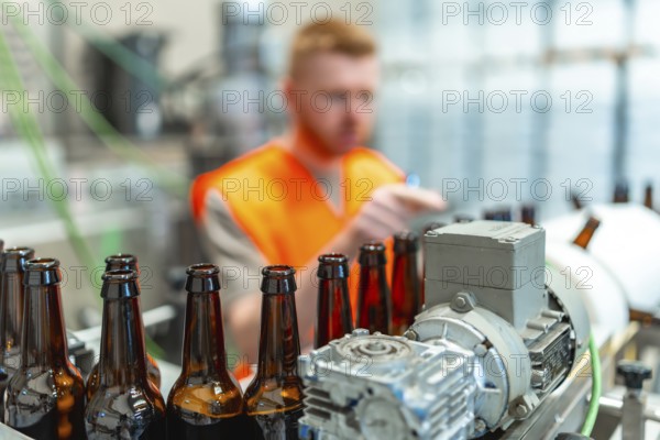 Brown glass bottles moving along a conveyor belt, with a factory worker in an orange vest monitoring the automated bottling process in a beer production plant