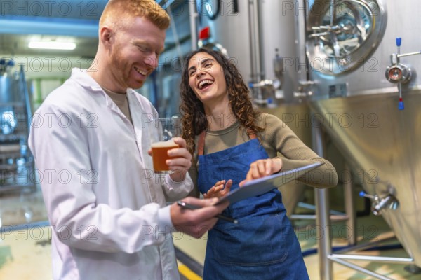 Man and woman tasting and checking craft beer quality, smiling and laughing together while discussing production data in a modern brewery or beer factory