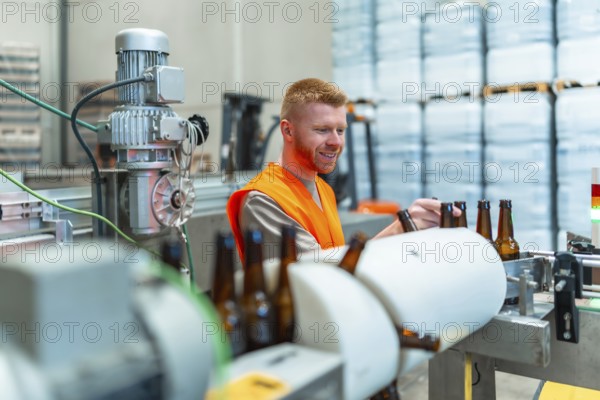 Smiling man in safety vest stands beside a conveyor belt, carefully inspecting brown glass bottles during the bottling process at a modern brewery factory