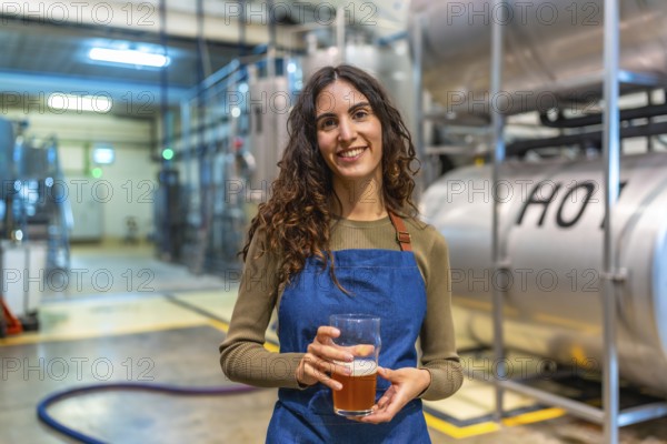 Woman brewer standing in modern brewery, smiling and holding a glass of craft beer for quality tasting, representing small batch production, craftsmanship and confident ownership