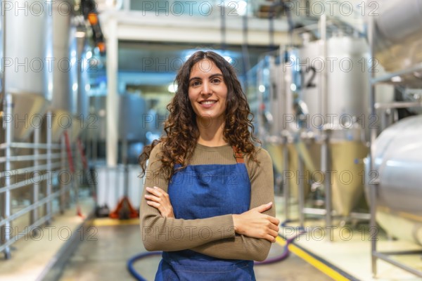 Woman brewer stands confidently with arms crossed, smiling at camera in a modern microbrewery with stainless steel fermentation tanks, proud small business owner and craftsperson